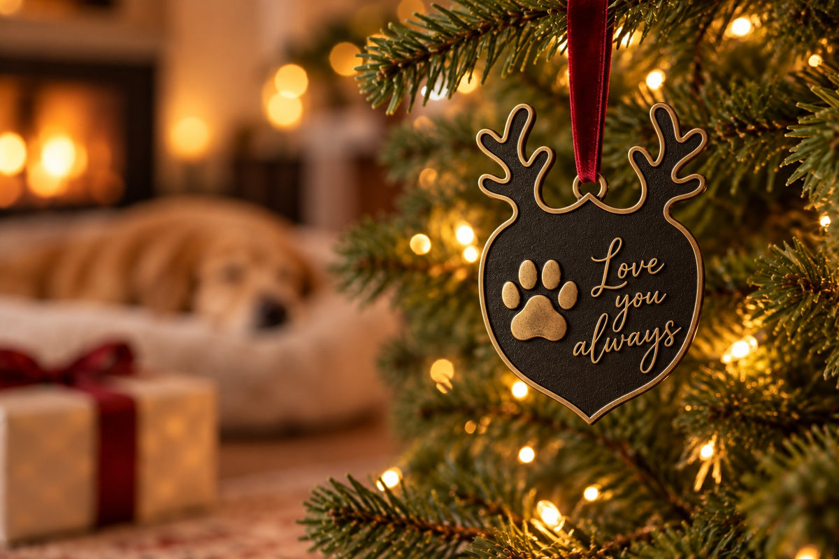 Antlered pet paw print ornament in antique brass reading Love you always, hanging by a red ribbon on a lit Christmas tree